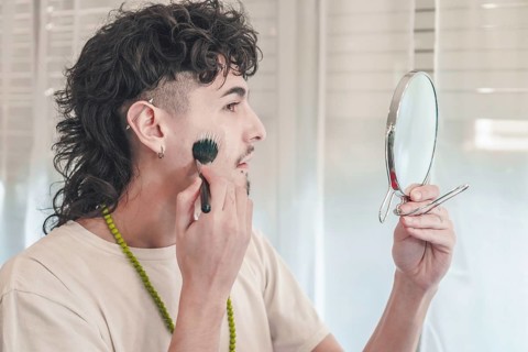 Young man with curly hair and piercings applying black makeup with a brush while holding a mirror,