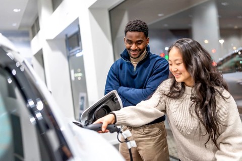 Happy multiethnic couple charging their new electric car at a dealership showroom, embracing a