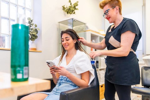 Hairdresser combing the hair of a client while she using mobile phone