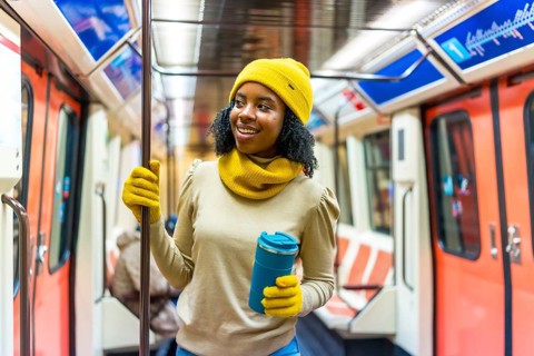 Smiling woman wearing yellow woolen hat and gloves holding a reusable mug while standing inside a