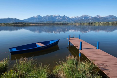 A lake with calm water and blue sky, a boat and a jetty in front of an alpine backdrop, Hopfen am