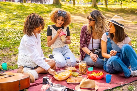 Four female friends are sitting on a blanket, eating snacks and drinking beverages during a picnic