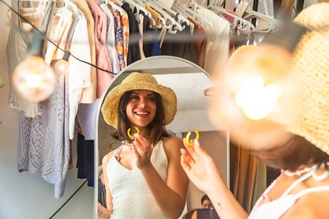 Young woman wearing straw hat trying on yellow earrings, getting ready in front of a mirror in her