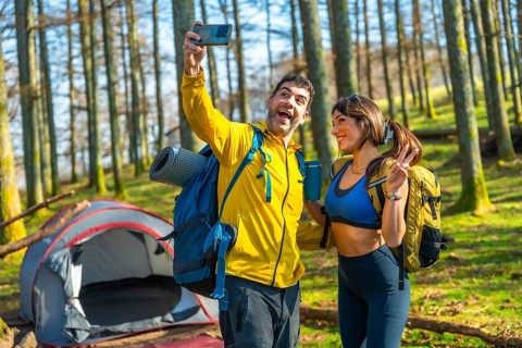Happy couple of hikers taking a selfie with smartphone in a forest next to their tent during a