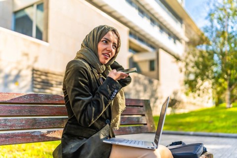 Arab young businesswoman sending voice message with mobile working with laptop sitting on a bench