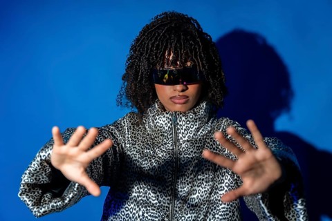 Young woman wearing futuristic visor and patterned jacket, gesturing with hands on blue background,