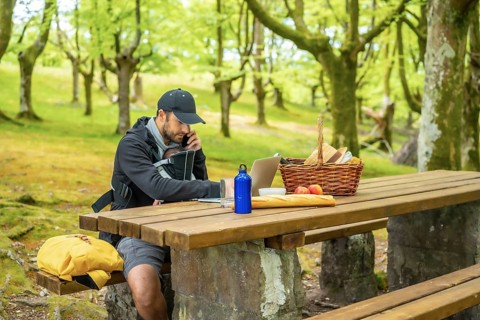 A young Caucasian father teleworking sitting at a picnic table with his computer and talking on the