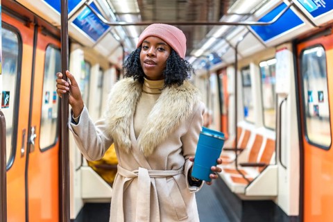 Stylish young black woman wearing winter coat and pink beanie holding reusable coffee cup while