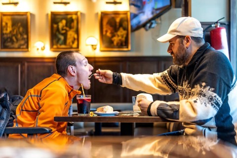 Man feeds his disabled friend in a restaurant, demonstrating care, friendship, and inclusion