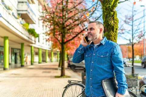 Mature man in denim clothes talking to the mobile and carrying laptop in the city in autumn