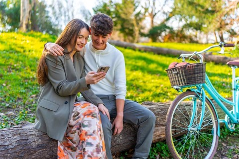 Smiling young couple sitting on a log in a park, sharing a moment on a smartphone, with a vintage
