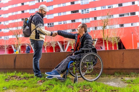 Tourist with backpack shaking hands with a disabled man in a wheelchair in a city park, with a red