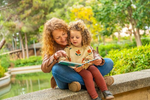 School girl sitting on the lap of her mother reading a book in a park