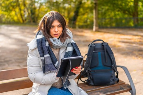 Woman with warm clothes working using digital tablet sitting on a park bench during sunny day of