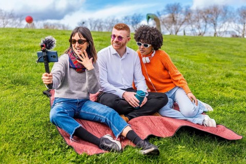 Three friends are recording a video blog in a park, using a smartphone with external microphone