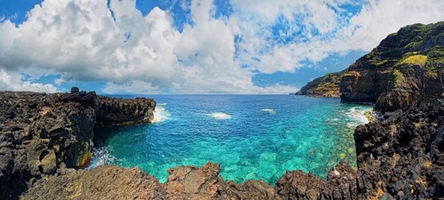 Panoramic view of a rocky lava coast with clear blue water and clouds in the sky, Termas da