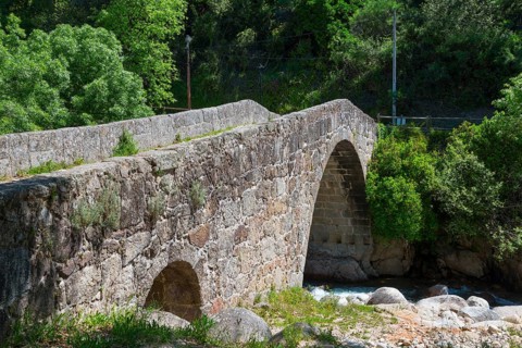 Old stone bridge over a river in a green, wooded setting under a sunny sky, Puente Parral, Roman