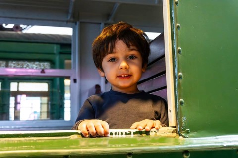 A young boy is sitting in a train car with his hands on the window. He is smiling and looking out