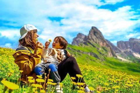 Mother and son playing with dandelions in a field of wildflowers with the seceda mountains in the