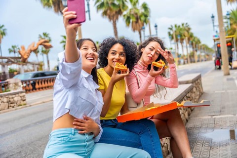 Multi-ethnic female friends taking a selfie while sharing pizza sitting in the street