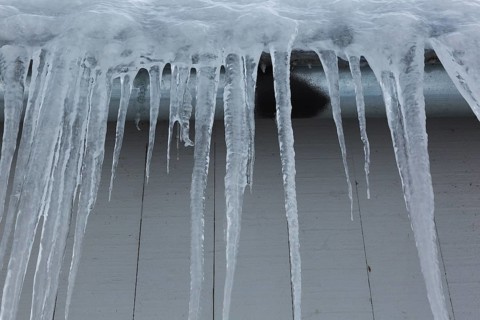 Long icicles and thick heavy ice hanging from roof edge of grey wood plank cladded building in