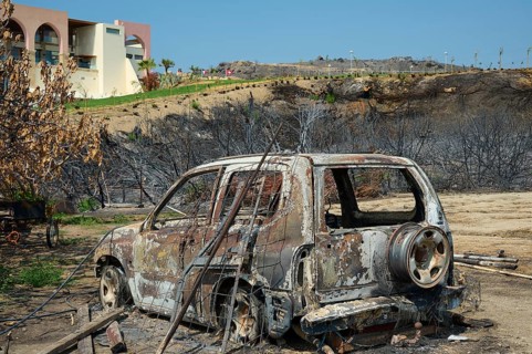 A burnt-out car stands in a destroyed landscape, destroyed trees and a building in the background,