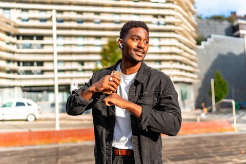African man listening to music using small wireless earbuds and dancing in the city