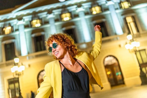 Happy woman in yellow jacket, black summer dress and sunglasses dancing alone in the city at night