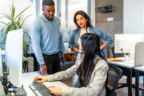 Office workers assisting a colleague using a computer and wearing a headset, possibly working in a