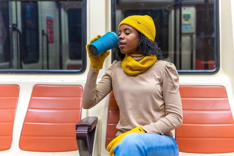 Young woman wearing winter clothes is sitting on a subway train, enjoying a warm beverage from her