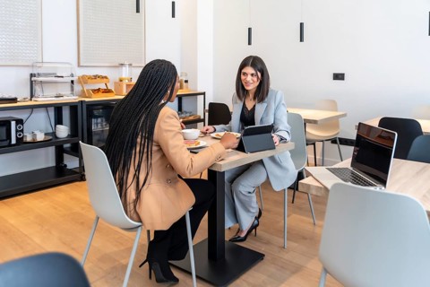 Two businesswomen enjoying breakfast and discussing work using a digital tablet in a modern hotel
