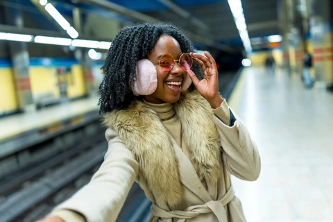 Smiling young woman wearing earmuffs and stylish glasses taking a selfie while waiting for the
