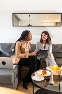 Two businesswomen are sitting on a sofa in a hotel room, using a tablet and laptop during a