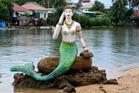 Greeting mermaid statue with white upper body and green tail fin sits on a stone on the shore,