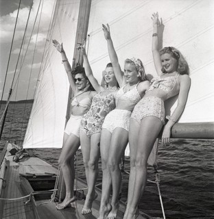 1940s sailing boat. Four young women are standing and leaning against the sails of the boat waiving