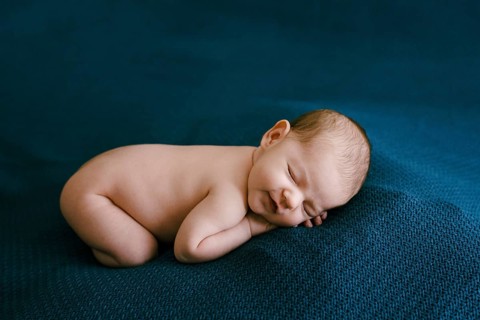 Naked newborn sleeping on a blue blanket with arms and legs tucked in.