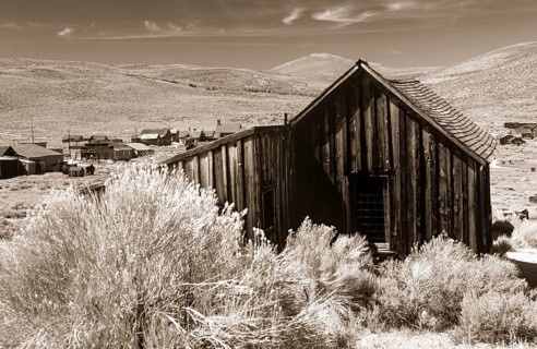 Old wooden shed in Bodie, behind it grassy hills of the prairie. Photo in sepia tone. Old wooden