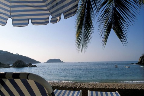 A tranquil beach scene with palm leaves, a striped umbrella, and a view of the ocean and distant