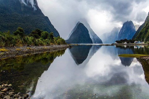 Mirror-smooth water of the Milford Sound fjord reflects mountains. Sunlight breaks through storm