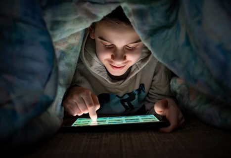Child watching his tablet in the bed. Illuminated child face from device screen. Smiling Boy under