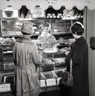 1960s coffee shop. Two women are standing in front of the counter that is full with freshly baked