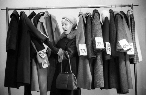 Great discounts of outerwear in the 1960s. A woman stands by a clothes rack, choosing from