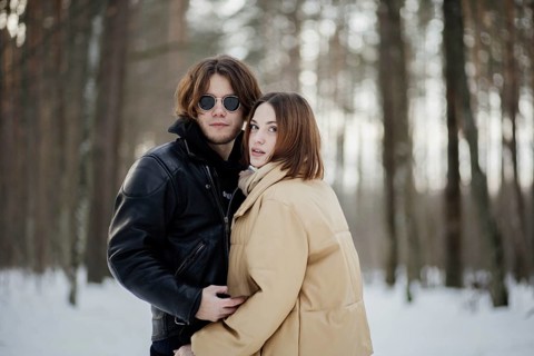 Couple poses in a snowy forest, looking at camera, dressed in winter jackets and scarves