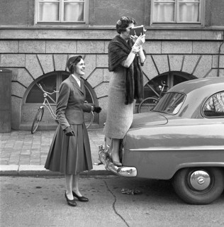 In the 1950s. An elderly woman with a film camera has stood on the bumper of a car to get a better