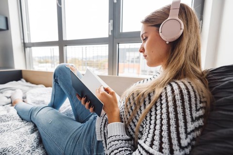 Blonde young caucasian woman lying on her dorm room bed next to a window putting on headphones,