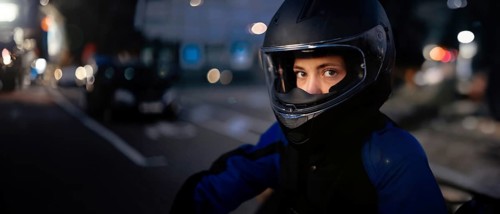 A person in a helmet and motorcycle gear standing by the street with urban lights in the background