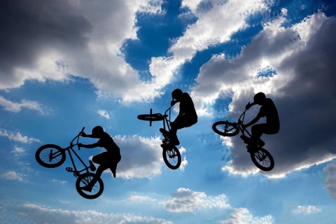 An extreme rider is making a free style jump from a ramp. The young boy with his bicycle is seen as