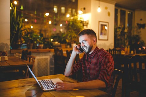 Young handsome Caucasian man with beard and toothy smile in red shirt works behind laptop, hands on