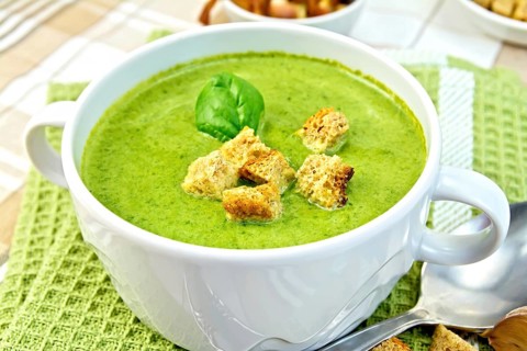 Green soup puree in a white bowl with croutons and spinach leaves on a napkin on the background