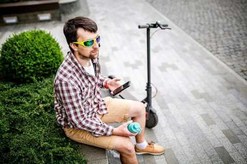 Man with e-scooter relaxing and using smartphone on bench in city. Modern male with electro scooter